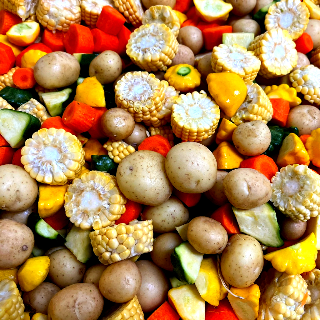 The image shows a colorful assortment of rustic vegetables. The mix includes whole baby potatoes, carrot chunks, zucchini pieces, yellow pattypan squash, and slices of corn on the cob. The vegetables are vibrant and varied, showcasing a range of colors from the earthy tones of the potatoes to the bright orange of the carrots, the green of the zucchini, and the pale yellow of the corn. The arrangement is dense, highlighting the freshness and variety of the vegetables.